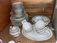Stack of white porcelain plates with gold trim, cups, sugar bowl with lid, and serving platters arranged on a shelf.