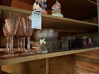 Full view of wooden shelf holding etched pink glass stemware, clear glass bowls, plates in clear and purple glass, and a small glass bell ornament with a green bow.
