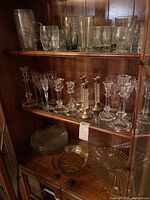 Full cabinet with upper shelves holding assorted glassware including tall glasses, mugs, clear crystal candleholders, and glass dishes on the bottom shelf.