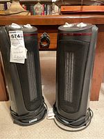 Two black cylindrical Everstar space heaters side by side on a carpet, with power cords wrapped at base, set in front of wood furniture.
