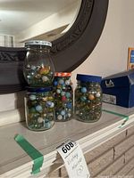 Three glass jars with screw-top lids filled with assorted vintage marbles, showing variety in size and color, placed on a white shelf with a mirror in the background.