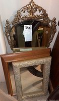 Four decorative wall mirrors stacked against a wall, featuring various carved and ornamental frames including a large curved gold-tone scrollwork frame and a rectangular wood frame.