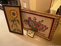 Three framed petit point needlepoint art pieces standing against wall on carpet floor: sunflower, floral bouquet, and cats.