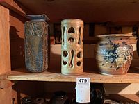 Three pottery vases on a wooden shelf: rectangular blue-toned vase, beige cylindrical vase with cut-outs, and rounded vase with brown glaze splashes.
