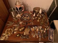 Full overview of the lot showing steins, souvenir spoons, brass figures, bells, and candle holders on a wooden table.