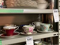 Shelf photo displaying six different tea cups and matching saucers with various floral and gold-trimmed designs.