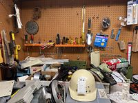 Photo showing a busy workbench area with a white hard hat, various tools, duct tape, electrical supplies, and other hardware items scattered on the work surface under a pegboard with tools.