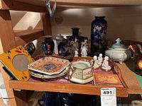 Shelf showing assortment of vases, plates, figurines, and a covered dish with pink cloth underneath.