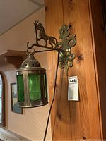 Photo showing the wall mounted lantern light with green glass panels hanging from a metal bracket featuring a horse figure and floral wall mount.