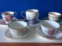 Five vintage cups and saucers arranged on a white surface against a blue background showing floral and gold trim designs.