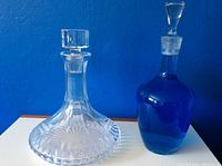 Photo showing both decanters side by side, the clear cut glass decanter on the left and the blue glass decanter on the right against a blue wall background.