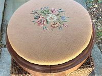 Top view of round footstool showing detailed embroidered floral pattern on beige fabric.