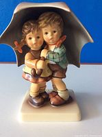 Front view of Hummel Sunshower figurine showing two children under umbrella, mounted on cream base.