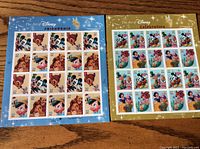 Two sheets of Disney postage stamps side by side on a wooden surface, showing the full sheets with the respective blue and gold backgrounds.