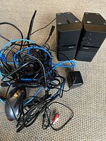 Pair of Bose speakers with black fabric covers showing volume and tone control knobs together with a bundle of wires and power adapter on a carpeted floor.