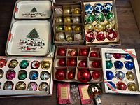 Five sets of vintage glass Christmas bulbs in original boxes, assorted colors and designs, plus three metal serving trays with holiday scenes on wood flooring.