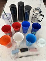 Overview of all items included in the lot: French press, three stainless steel bottles, Starbucks tumbler, seven reusable plastic cups, three reusable fold-up straws and cases, and a white plastic lid.