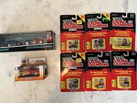 Photo showing a boxed 1932 Ford Roadster with traveler trailer by Maisto, an M2 Machines Ground Pounders red truck in blister pack, and six Racing Champions 1992 1:64 scale die-cast cars in sealed packaging.