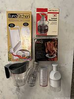 Photo showing box of EuroKitchen Professional Chef's Slicer, One Touch Instant Marinator box, Chicken Roaster box, and loose kitchen containers: salad dressing bottle, measuring container, and clear plastic bottle with black handle.