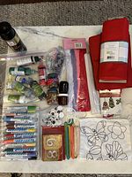 Overview of craft supplies displayed on table including fabric markers, paint bottles, red felt fabric, craft ornaments, googly eyes, sequins, colored pipe cleaners, and butterfly-print backpack.