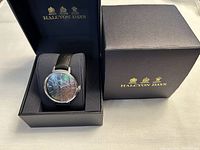 Halcyon Days ladies watch displayed inside a black Halcyon Days branded box with gold logos, angled view showing watch face and strap, black dial with subtle pattern and silver-tone bezel.