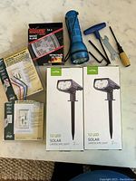 Photo showing two boxes of Litom 12 LED solar landscape lights, blue flashlight, First Alert smoke alarm, fan control, and assorted tools.