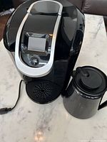 Photo showing the Keurig coffee machine with digital display and button, alongside the stainless steel family size coffee pot with lid on white marble surface.