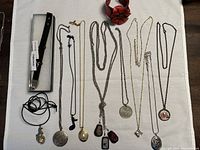 Photo showing variety of necklaces and two bracelets arranged on cloth surface. Includes different chain styles and pendants such as lockets, dog tags, floral and crystal accents.