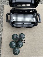 Top view of portable BBQ with open lid and cooking grate, alongside five green propane canisters arranged in a cluster on a woven mat