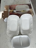 Two white rectangular ceramic serving dishes with grapevine embossed borders shown outside box; original box in background