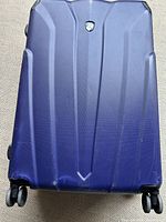Front view of a large blue hard cover suitcase showing spinner wheels and textured surface with a logo badge.