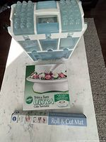 Photo of the Wilton large plastic storage/carry case in white and light blue compartments, Wilton Trim 'n Turn Ultra Cake Turntable box, and Roll & Cut Mat box placed on a marble surface.