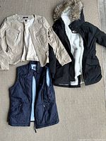 Three women’s outerwear items including beige leather jacket, black winter coat with fur hood and dark blue vest.