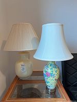 Two decorative table lamps side by side on a glass table against a wall, one with a floral yellow base and white shade, the other cream base with pleated shade.