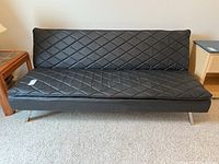 Front view of black vinyl futon showing diamond quilted pattern upholstery and chrome legs, placed on carpet next to wooden side tables.