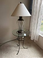 Full view of glass top side table with iron base and decorative glass lamp on top near window with curtains and carpeted floor.