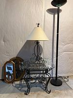 Full view of glass table with black iron base, table lamp with beige shade, tall floor lamp, and two wall mirrors leaning on the wall.