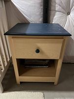 Photo of small pine nightstand with light pine body and black top surface, drawer with round knob and open lower shelf with items inside. Visible surface marks and wear on the black top.