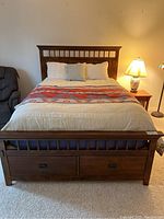 Front view of queen wooden bed frame with beige bedding and red patterned throw, showing headboard and footboard with under-bed drawers.