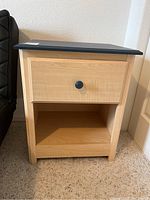 Front and side view of one wood side table with drawer and open shelf, natural wood finish with dark blue top, positioned next to a sofa on carpet floor, showing overall condition and design.