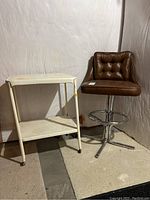 Brown vintage bar stool with padded, button-tufted vinyl seat and back on chrome adjustable base with footrest, next to white metal utility cart with two shelves.