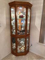 Front view of the tall wood corner curio cabinet, showcasing the glass doors and internal shelves, with some items inside to show scale and lighting.