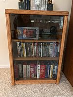 Front view of a wood media cabinet filled with DVDs behind glass doors. The cabinet is placed on a carpeted floor.