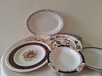 Seven antique decorative porcelain plates and platters in various floral and ornamental designs, arranged on a white surface