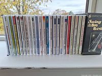 View of the spines of 17 CD cases showing a mix of classical, brass band, pop, and musical titles, arranged horizontally on a window ledge.