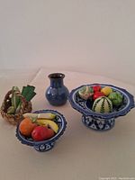 Two miniature blue and white ceramic pedestal dishes filled with realistic miniature fruit and vegetables, a small blue vase, and a basket with vegetables placed on a white surface