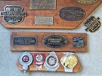 Photo showing multiple mounted brass and metal plates including Lancashire Dynamo & Motor Co, Laurie & Lamb Montreal, Pruston & Hornsby England, and automobile association badges.