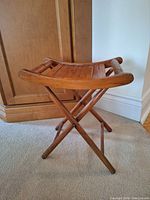 Side angle of the wooden folding artist's seat showing the slatted curved wooden seat and crossed leg construction.