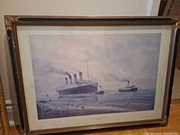 Framed print showing RMS Titanic leaving Belfast for sea trials, clear depiction of ship and water, antique style frame with visible wear.