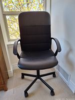 Front view of black executive office chair with armrests and wheeled base near a window.